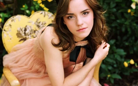 Emma Watson sitting on a yellow floral chair in a garden setting, wearing a peach dress with a black bow. The image is a high-definition desktop wallpaper and background.