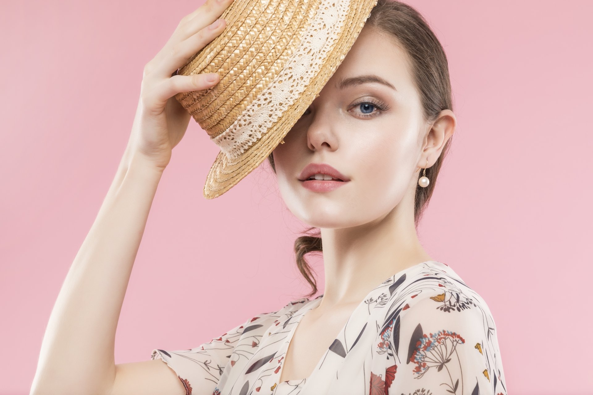 A woman with striking blue eyes lifts a straw hat, posing against a soft pink background in this 4K Ultra HD PC desktop wallpaper and model portrait.