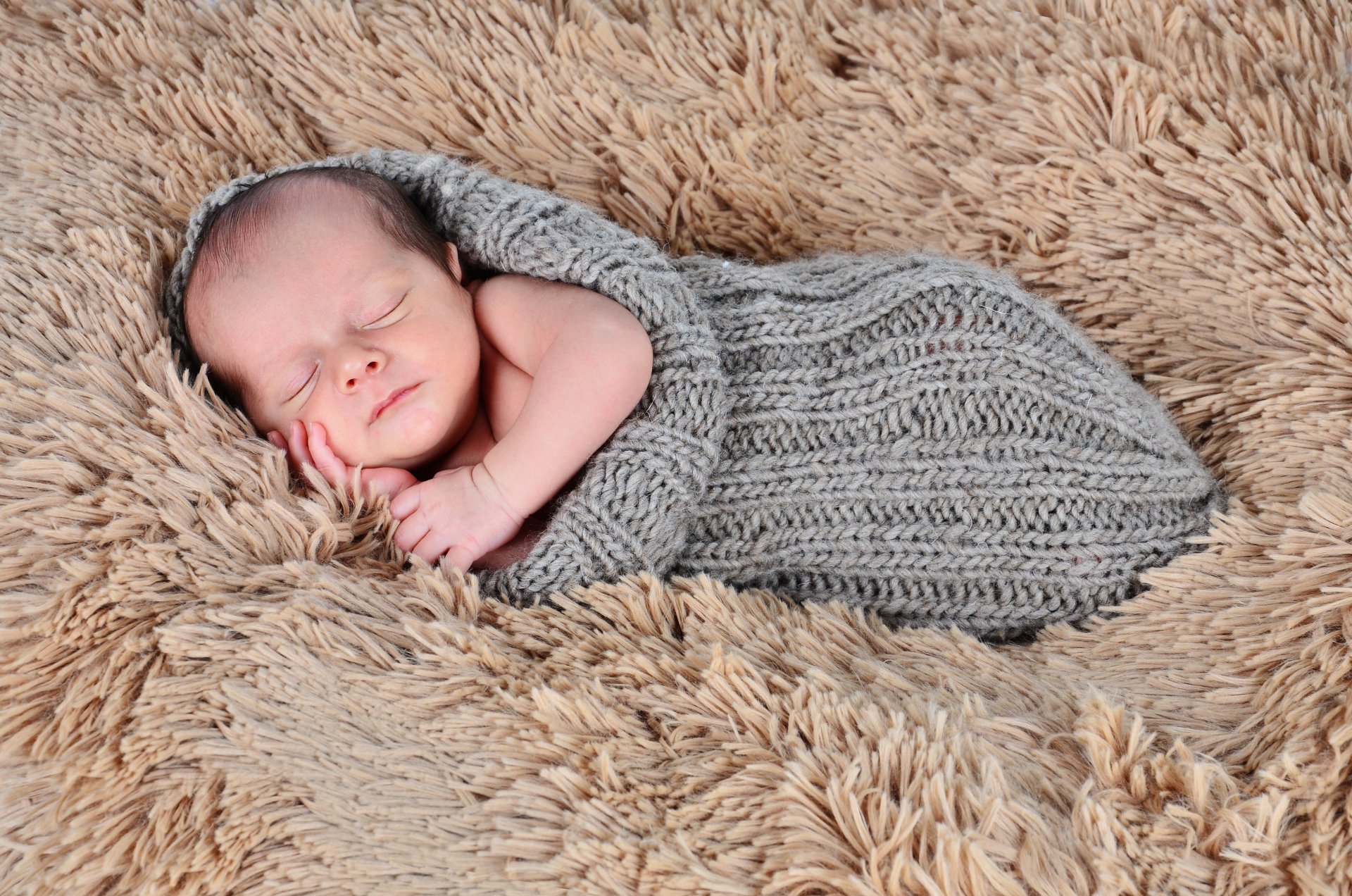 A cute baby peacefully sleeps wrapped in a cozy gray knit blanket, resting on a soft, textured fur surface. This 4K Ultra HD wallpaper captures a serene moment.