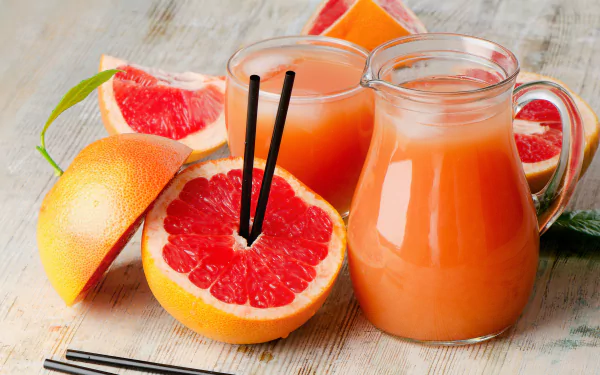 A vibrant 4K Ultra HD still life of fresh grapefruit, a glass of juice with two black straws, and a pitcher filled with grapefruit juice on a wooden surface.