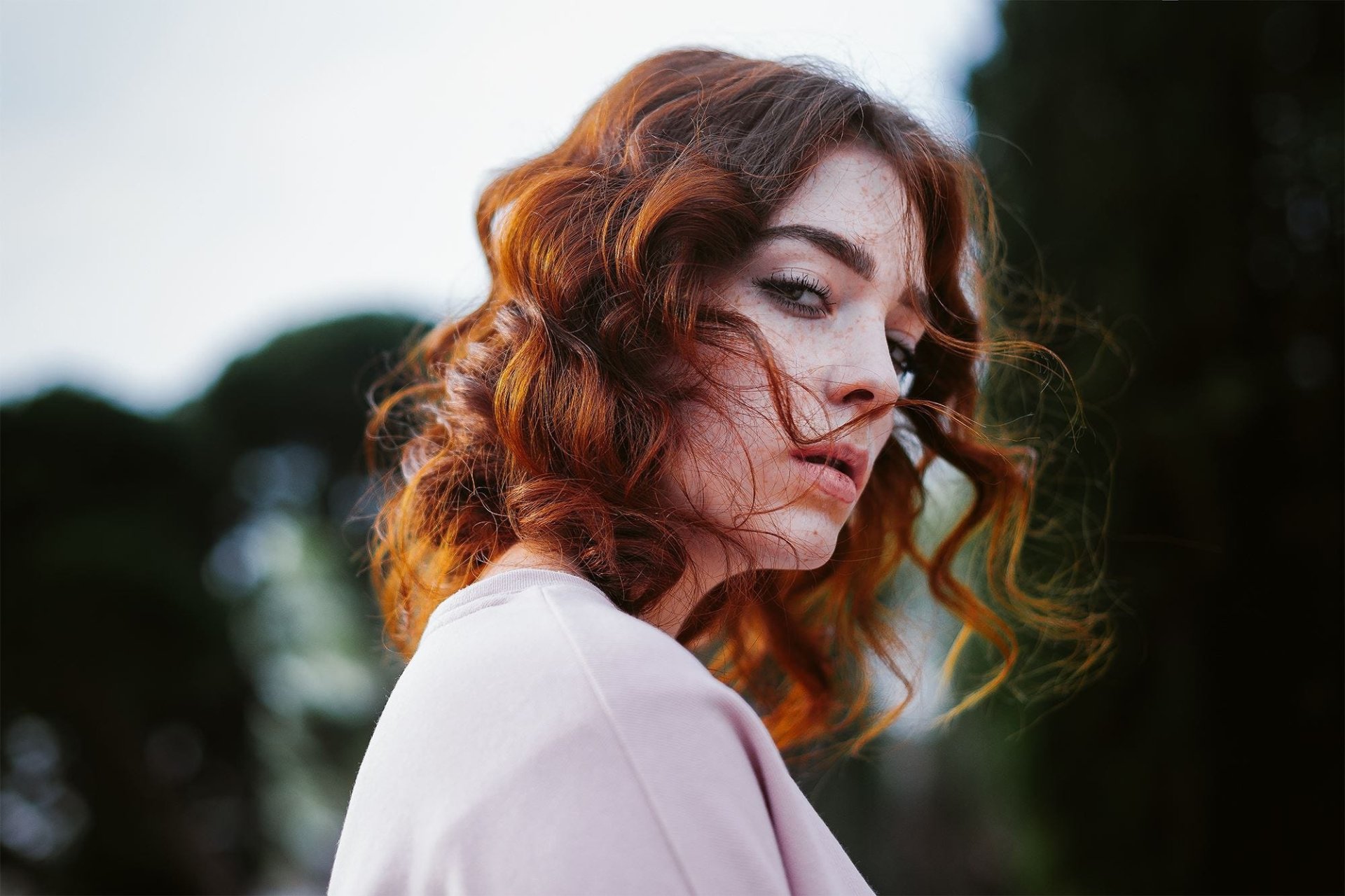 Close-up HD desktop wallpaper of a freckled redhead model gazing over her shoulder, wind-tousled curls and soft bokeh background.
