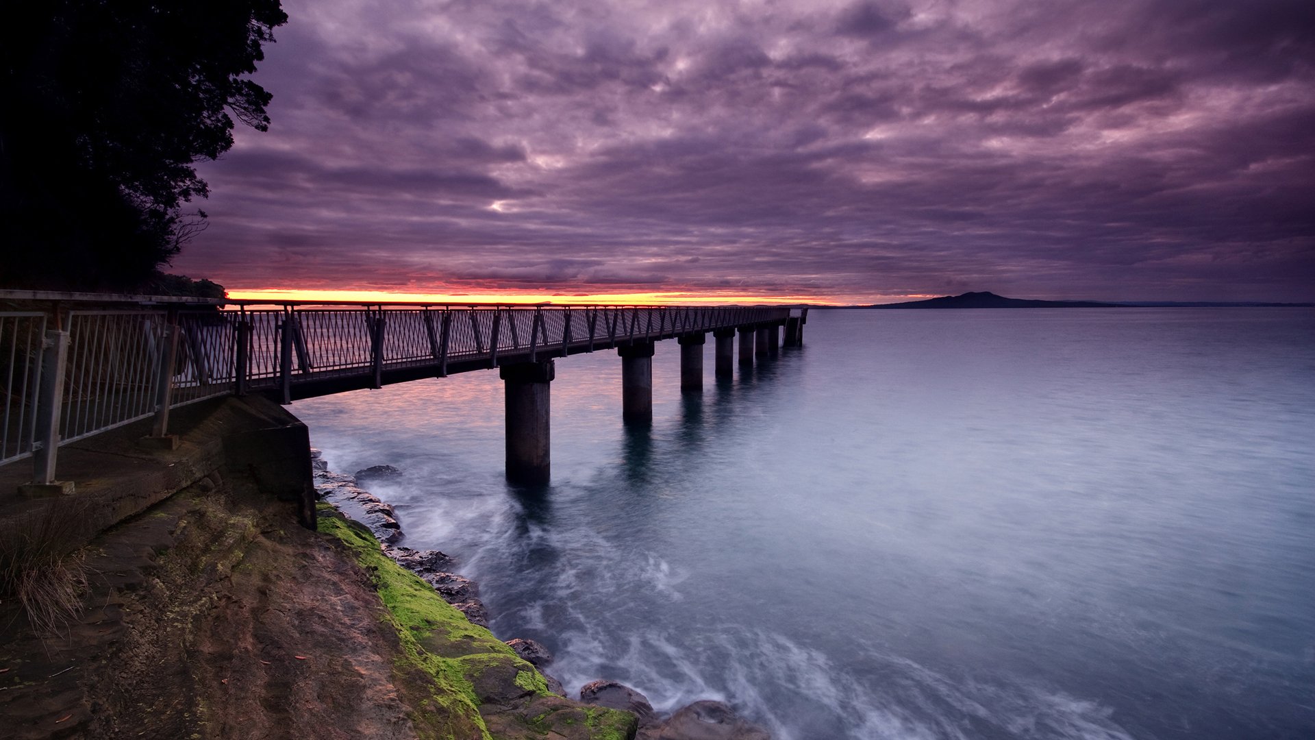A serene sunset over the ocean, with a dock extending into calm waters. The sky is filled with purple and orange hues, reflecting the beauty of nature along the pier.