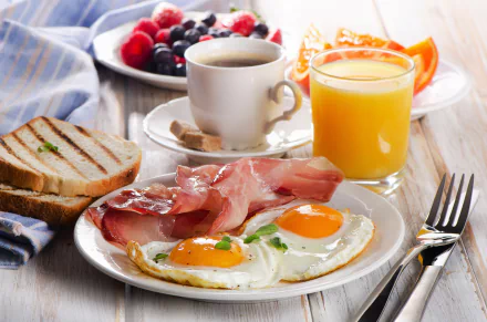 HD desktop wallpaper featuring a breakfast spread with toast, fried eggs, bacon, fresh fruit, coffee, and a glass of orange juice on a wooden table.
