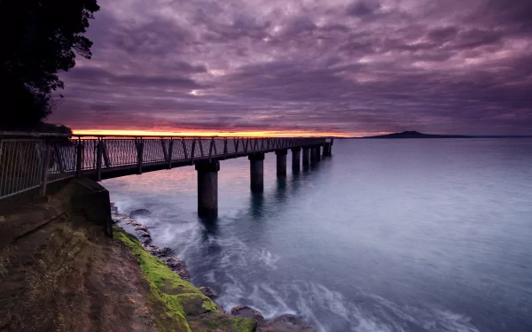 A serene sunset over the ocean, with a dock extending into calm waters. The sky is filled with purple and orange hues, reflecting the beauty of nature along the pier.