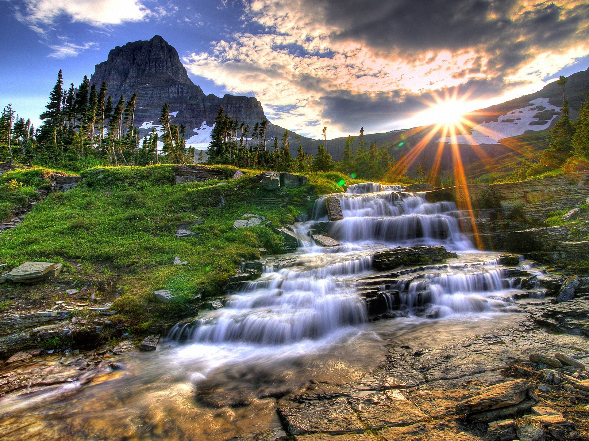 Cascading waterfall with sunlit mountain and stream in serene nature setting.