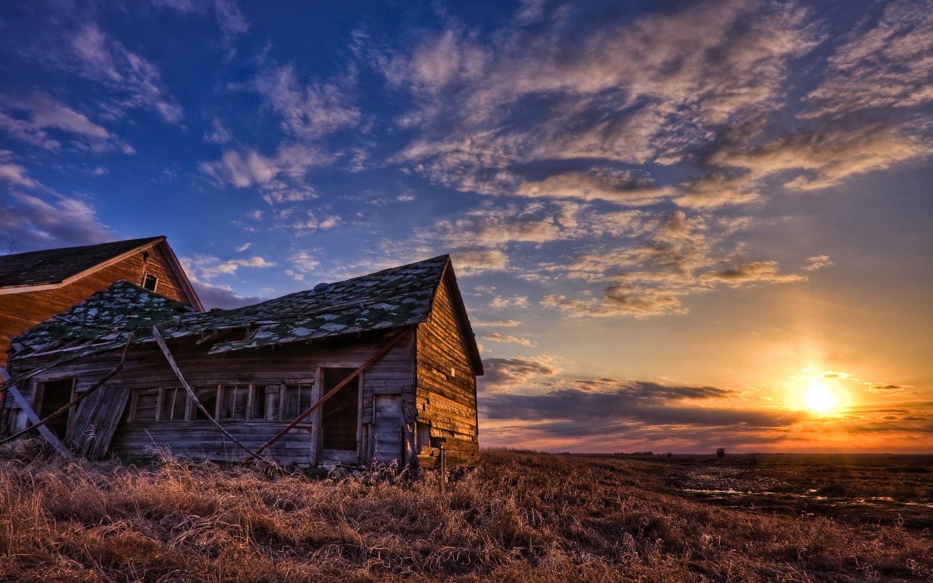 A rustic, man-made wooden building stands on earth under a vivid sky with the sun setting, creating an HD desktop wallpaper with warm and natural tones.