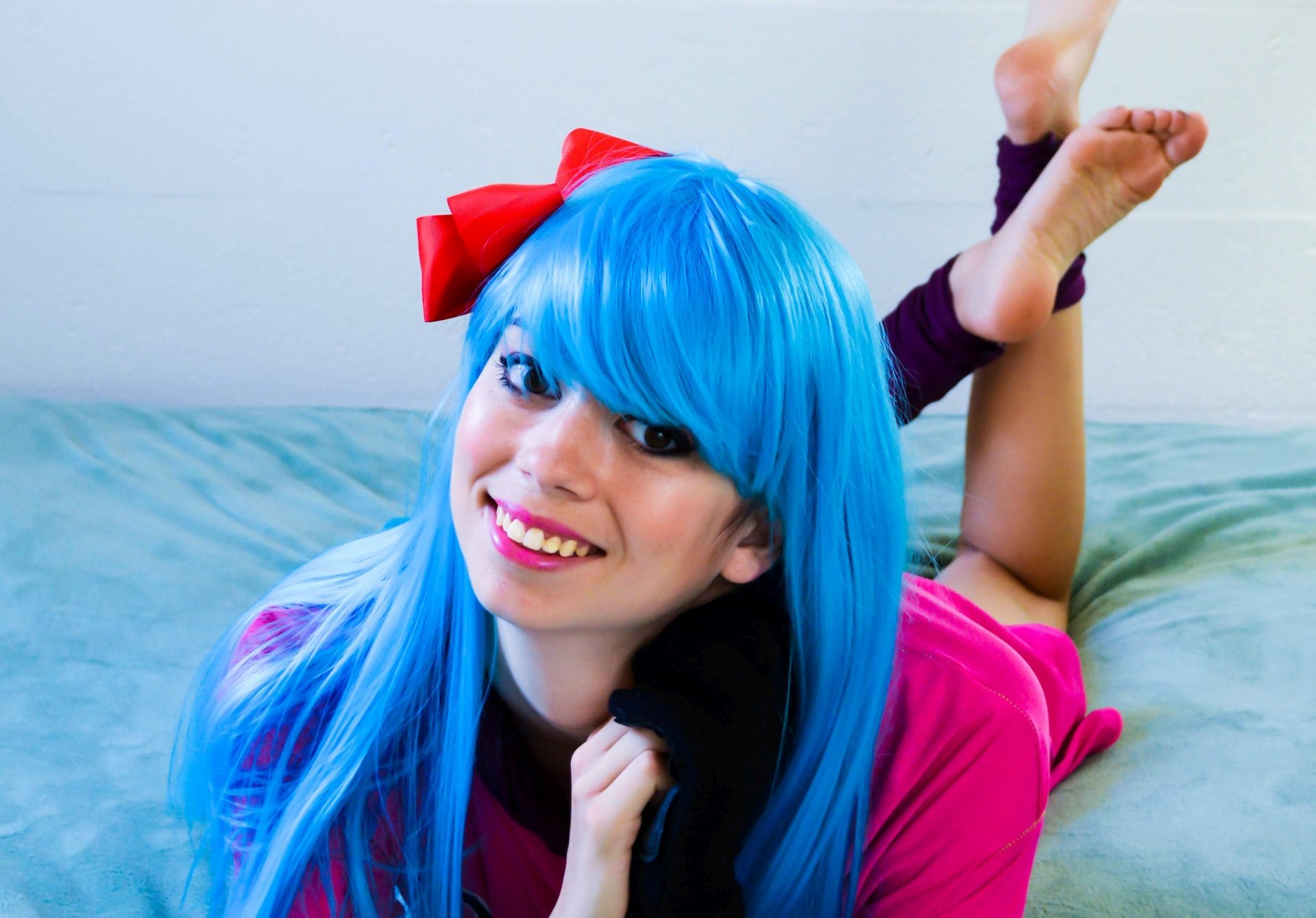 2K Quad HD PC desktop wallpaper of a woman cosplaying Bulma (Dragon Ball) — smiling in a blue wig, pink outfit and red bow, playful pose on a bed.