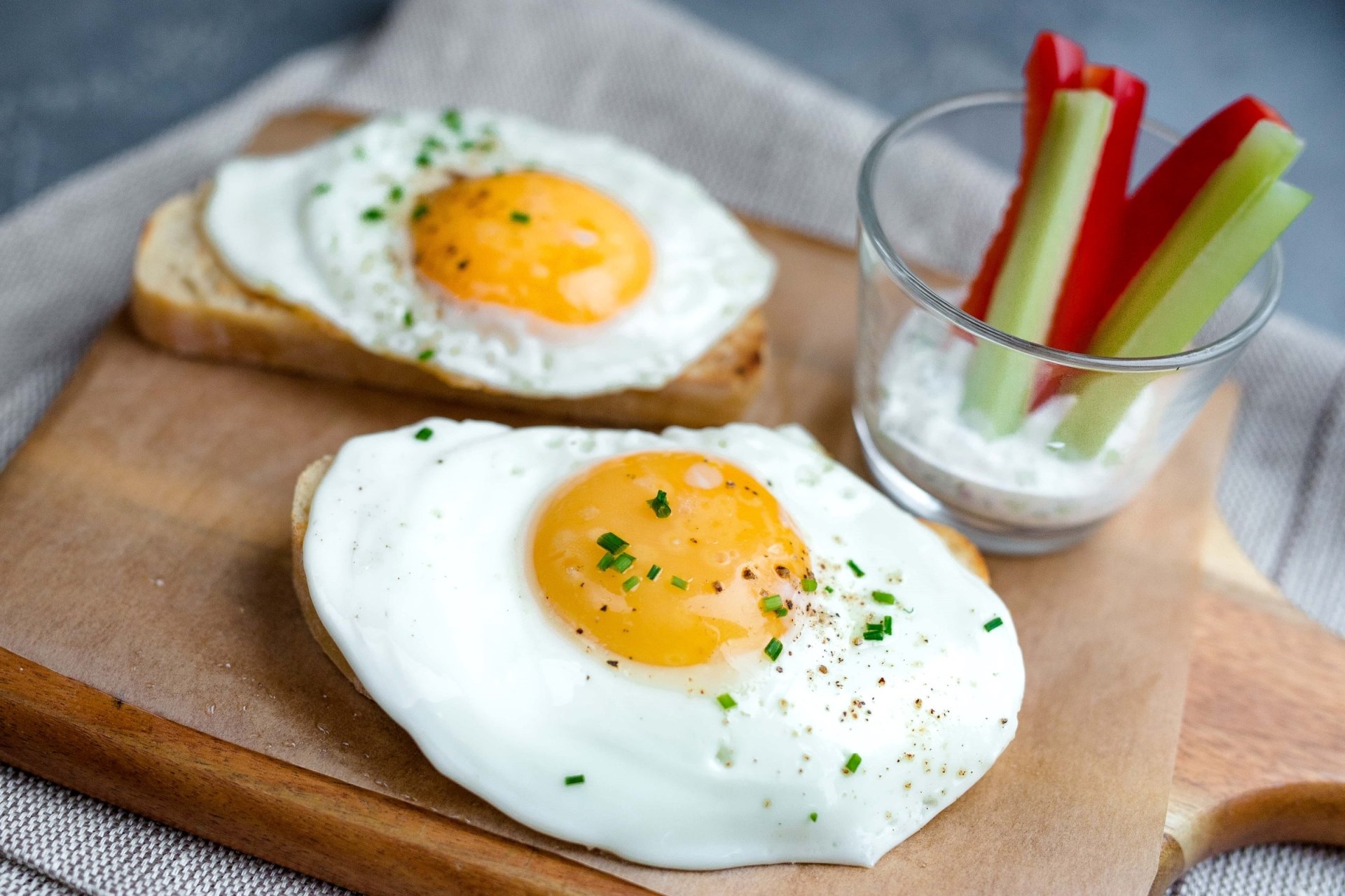 HD PC desktop wallpaper of breakfast food: two sunny-side-up eggs on toast on a wooden board, sprinkled with chives, beside a small glass of celery and red pepper sticks.