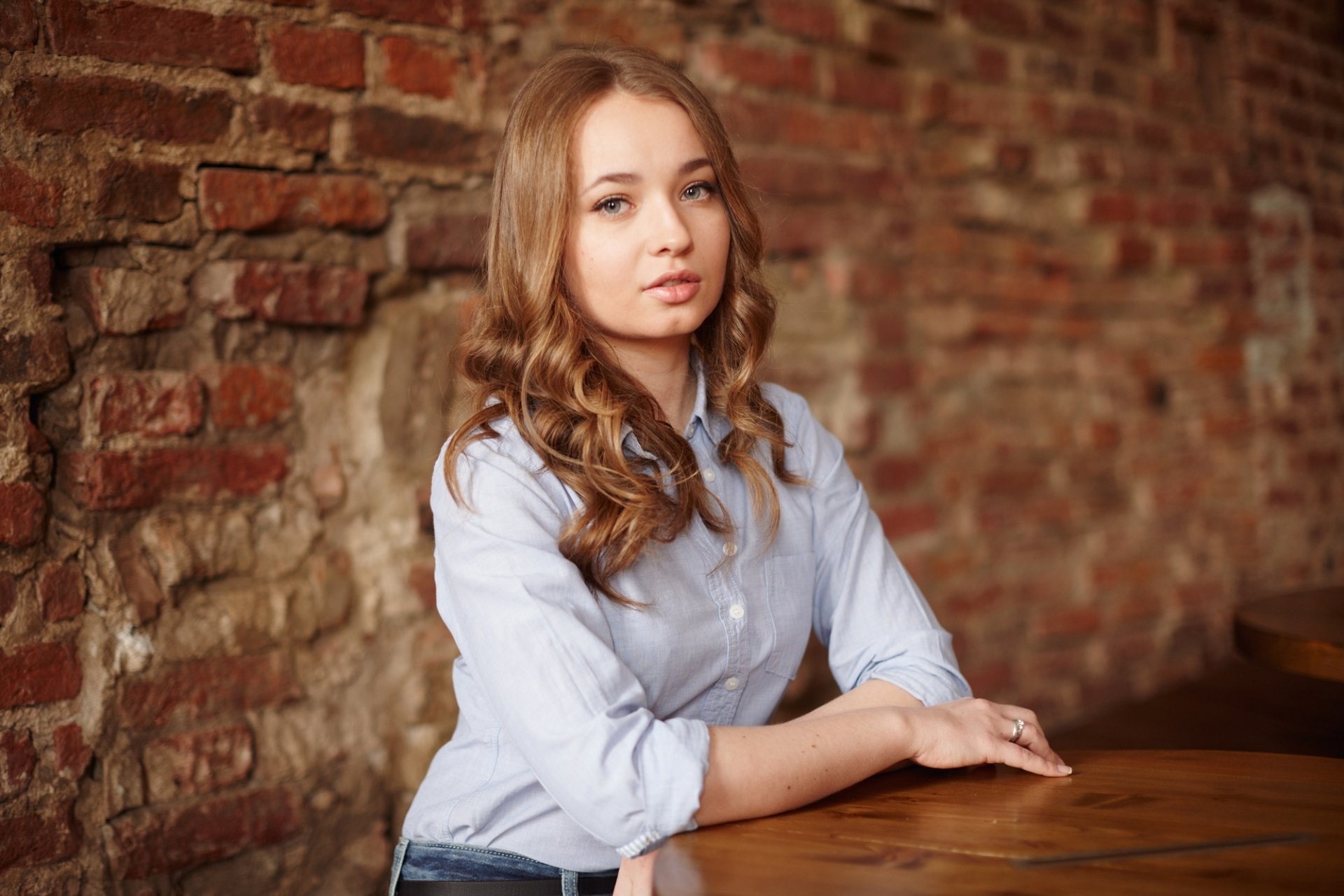 HD desktop wallpaper featuring a redhead woman with blue eyes and a calm expression, seated against a rustic brick wall background.
