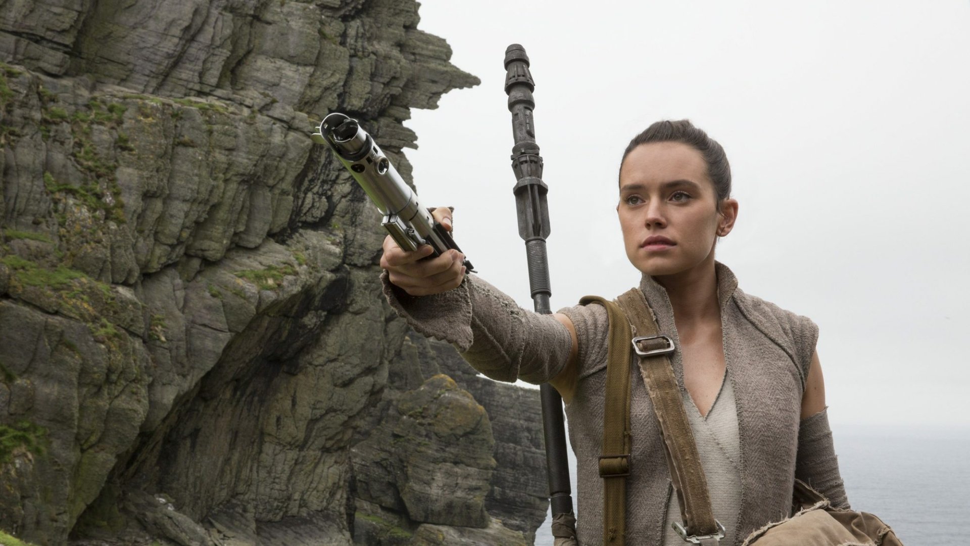 HD desktop wallpaper featuring Daisy Ridley as Rey from Star Wars: The Last Jedi. Rey is holding a lightsaber, positioned against a backdrop of rocky cliffs.