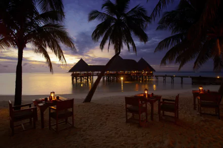 Evening view of a Maldives beach with palm trees, a bungalow on stilts over the water, and a few tables and chairs on the sand under a colorful horizon, captured in HD.