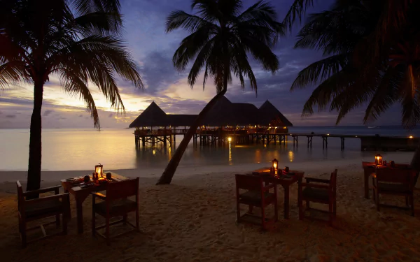Evening view of a Maldives beach with palm trees, a bungalow on stilts over the water, and a few tables and chairs on the sand under a colorful horizon, captured in HD.