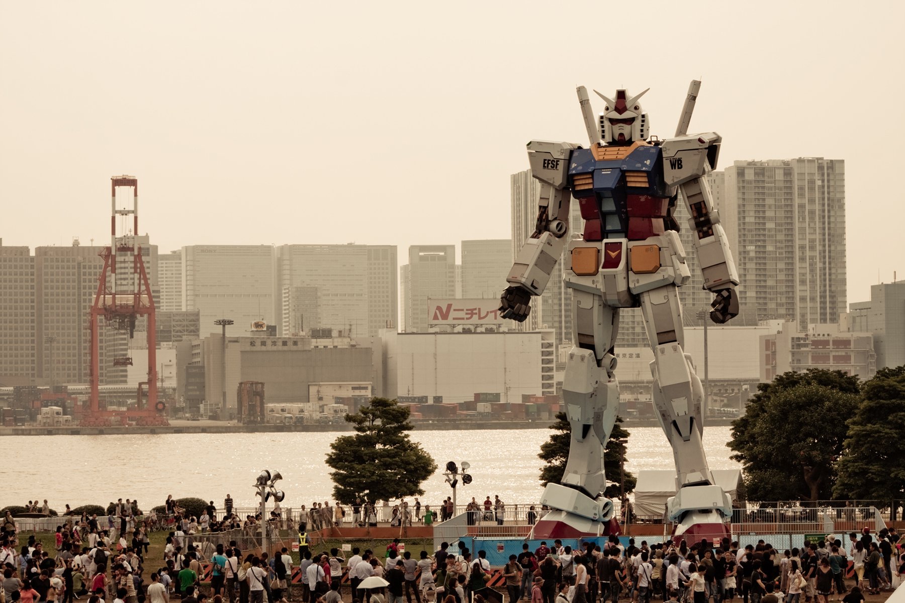 A stunning HD desktop wallpaper featuring a giant Gundam robot towering over a crowd by the water, with a city skyline in the background, capturing the essence of anime culture.