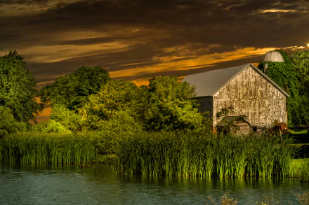A serene HD desktop wallpaper featuring a farm with a rustic barn by a river, set against a vibrant sunset sky with lush green surroundings.