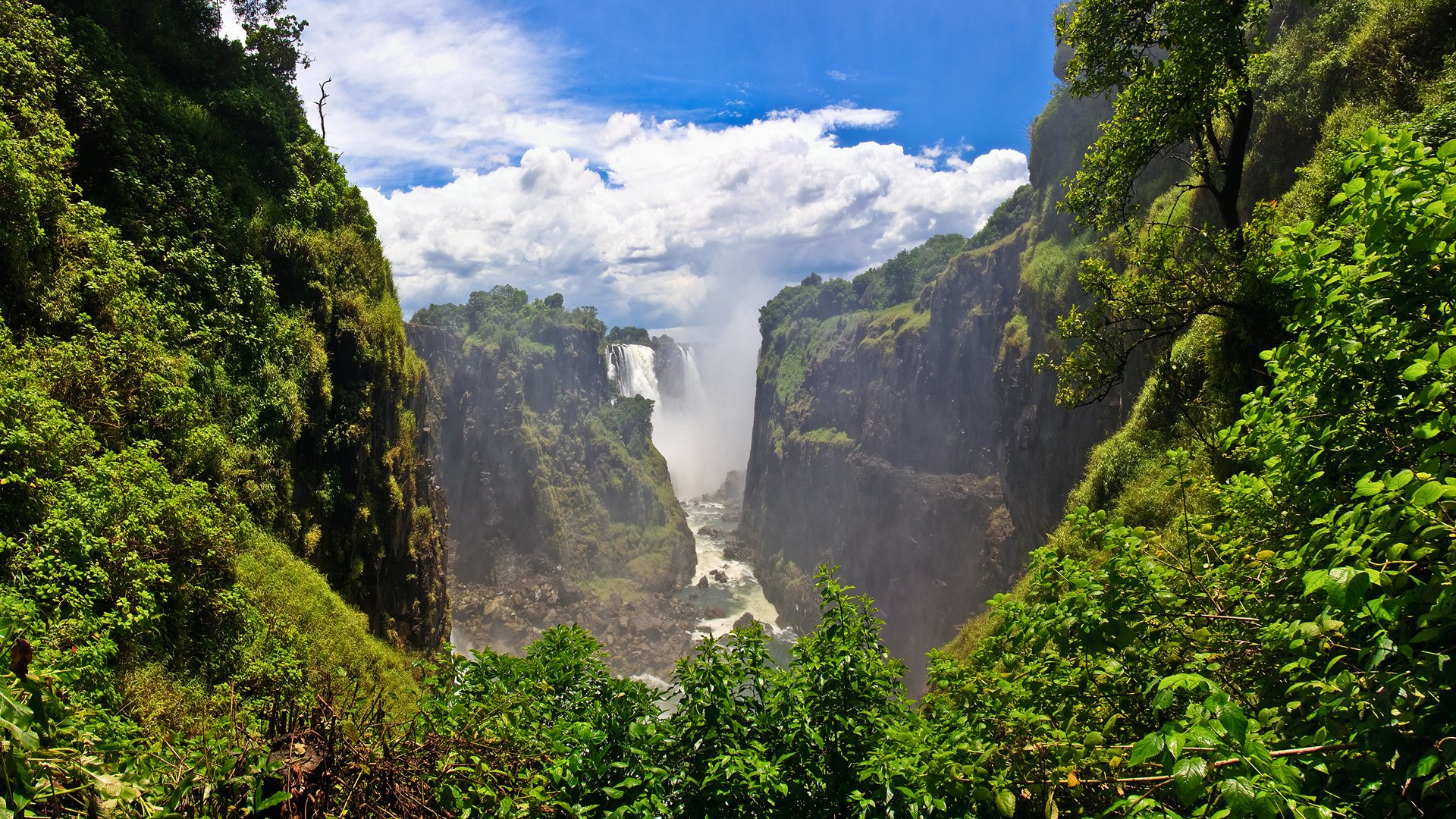 A stunning HD wallpaper showcasing a lush green landscape with a majestic waterfall cascading between cliffs, under a vibrant sky dotted with clouds. Nature's beauty captured in tranquil harmony.