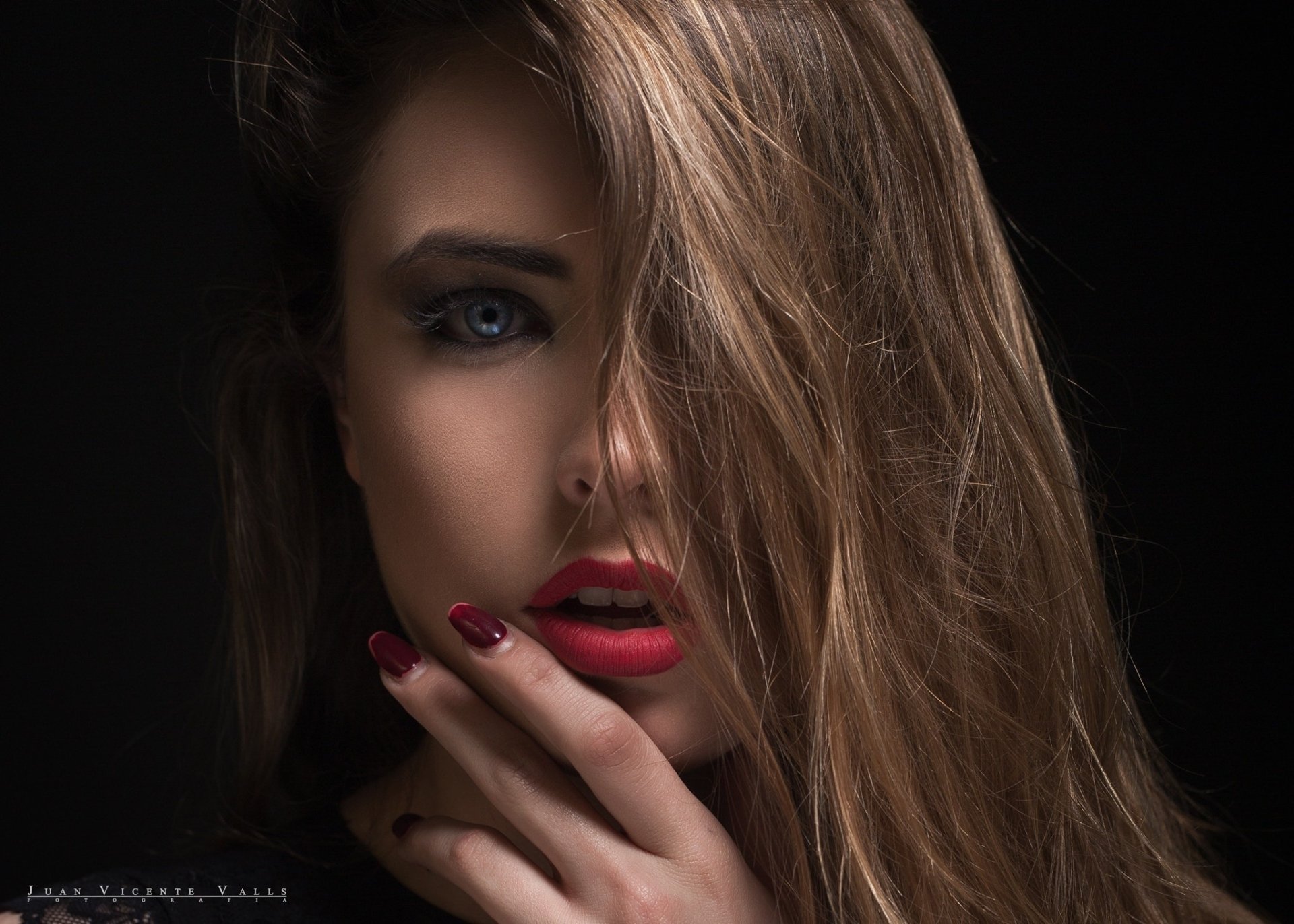A high-definition desktop wallpaper featuring a brunette model with blue eyes, wearing lipstick, with a captivating expression. The woman’s face is partly covered by her hair against a dark background.