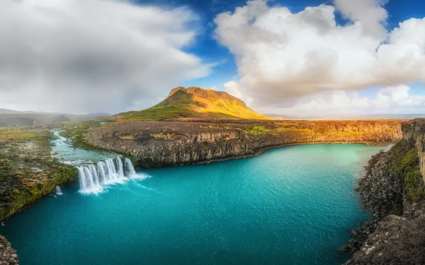 Stunning HD landscape of Iceland's Goðafoss waterfall cascading into a turquoise river, surrounded by rugged mountains and dramatic clouds.