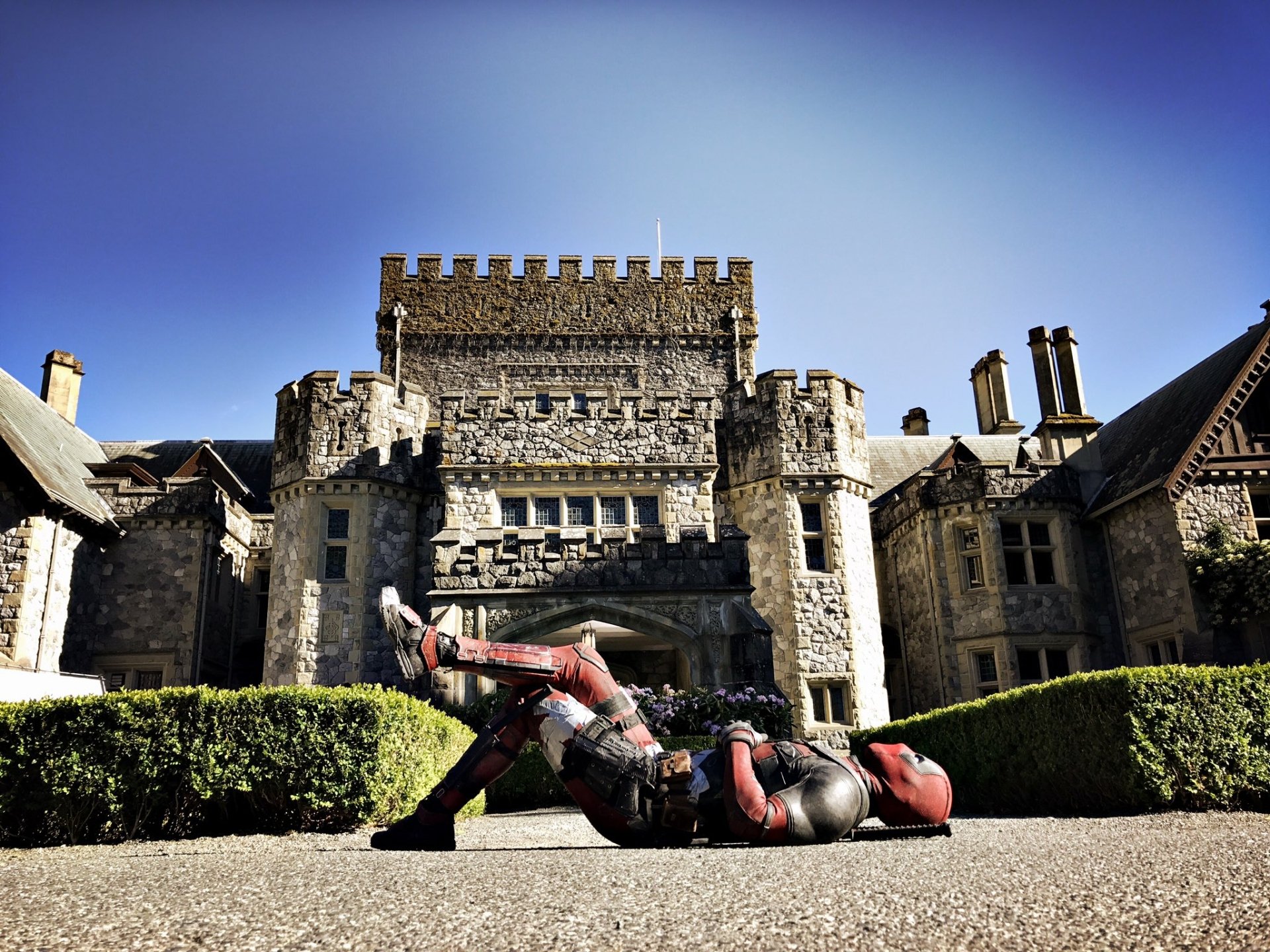 Deadpool sprawls on the ground in front of a grand stone castle, capturing a moment from the movie Deadpool 2. This vibrant HD image serves as an engaging desktop wallpaper.