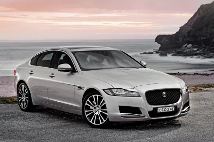 Silver Jaguar XF sedan parked by a rocky coastline under a serene sunset, captured in stunning 4K Ultra HD for PC desktop wallpaper and background.