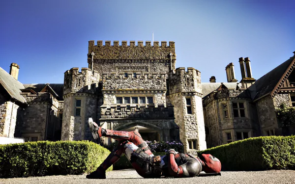 Deadpool sprawls on the ground in front of a grand stone castle, capturing a moment from the movie Deadpool 2. This vibrant HD image serves as an engaging desktop wallpaper.