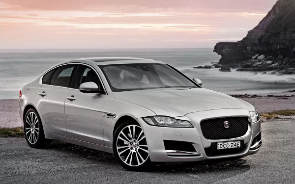Silver Jaguar XF sedan parked by a rocky coastline under a serene sunset, captured in stunning 4K Ultra HD for PC desktop wallpaper and background.