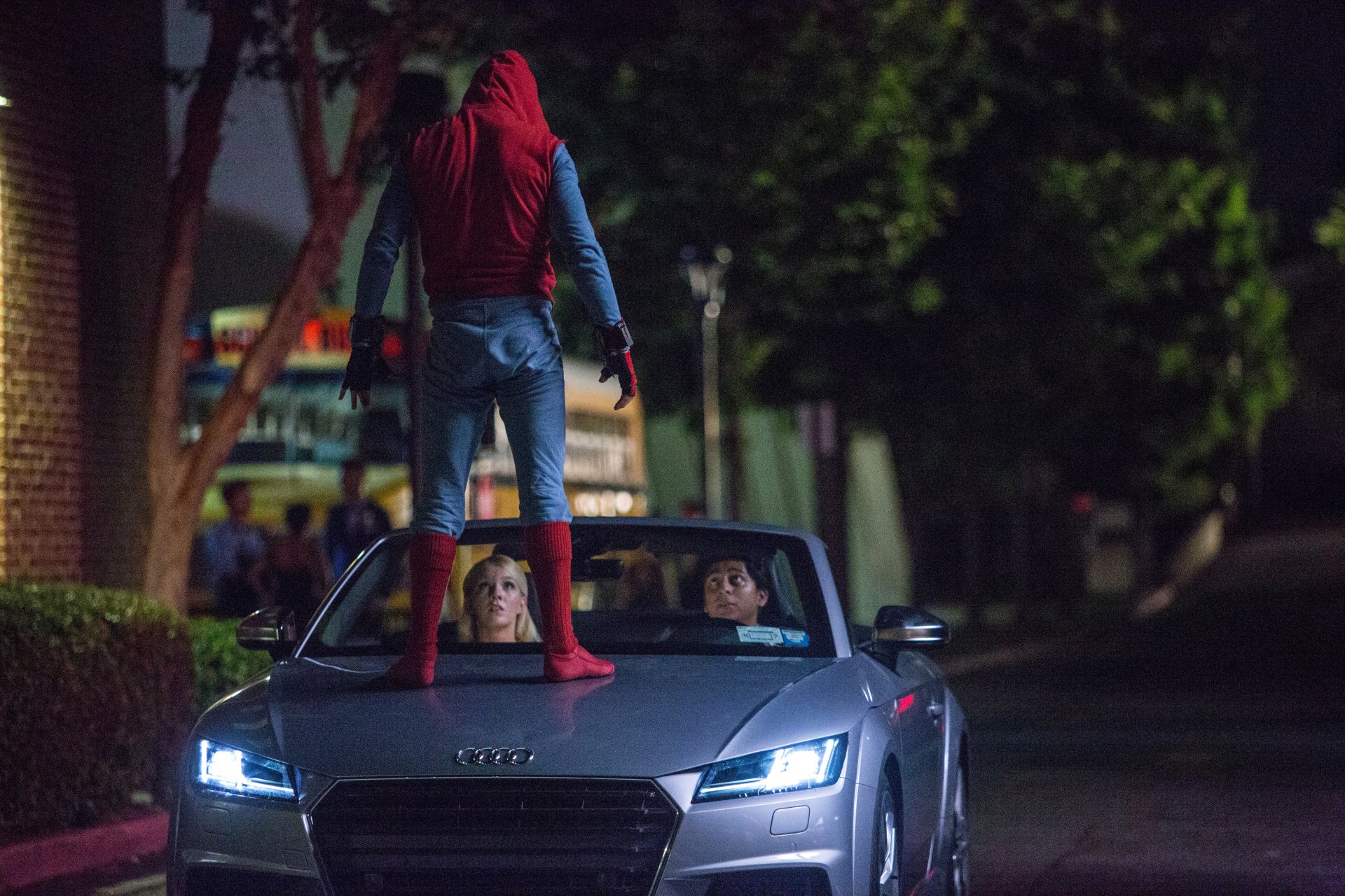 Spider-Man stands on the hood of a silver car at night, confronting two people inside. Scene from the movie Spider-Man: Homecoming. This HD desktop wallpaper and background captures the action and drama.