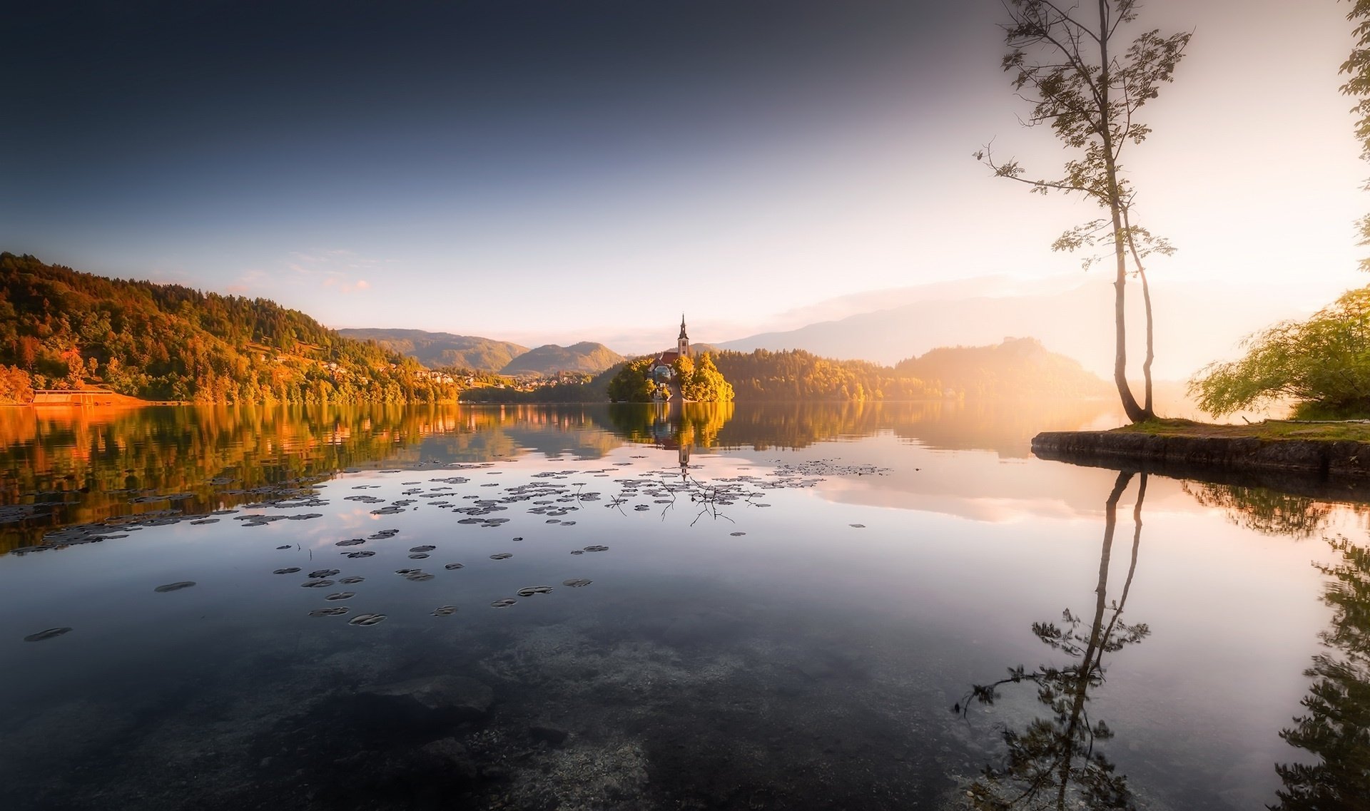 Serene view of the Assumption of Mary Church reflecting on Lake Bled at sunrise, framed by calm waters and distant autumn hills.