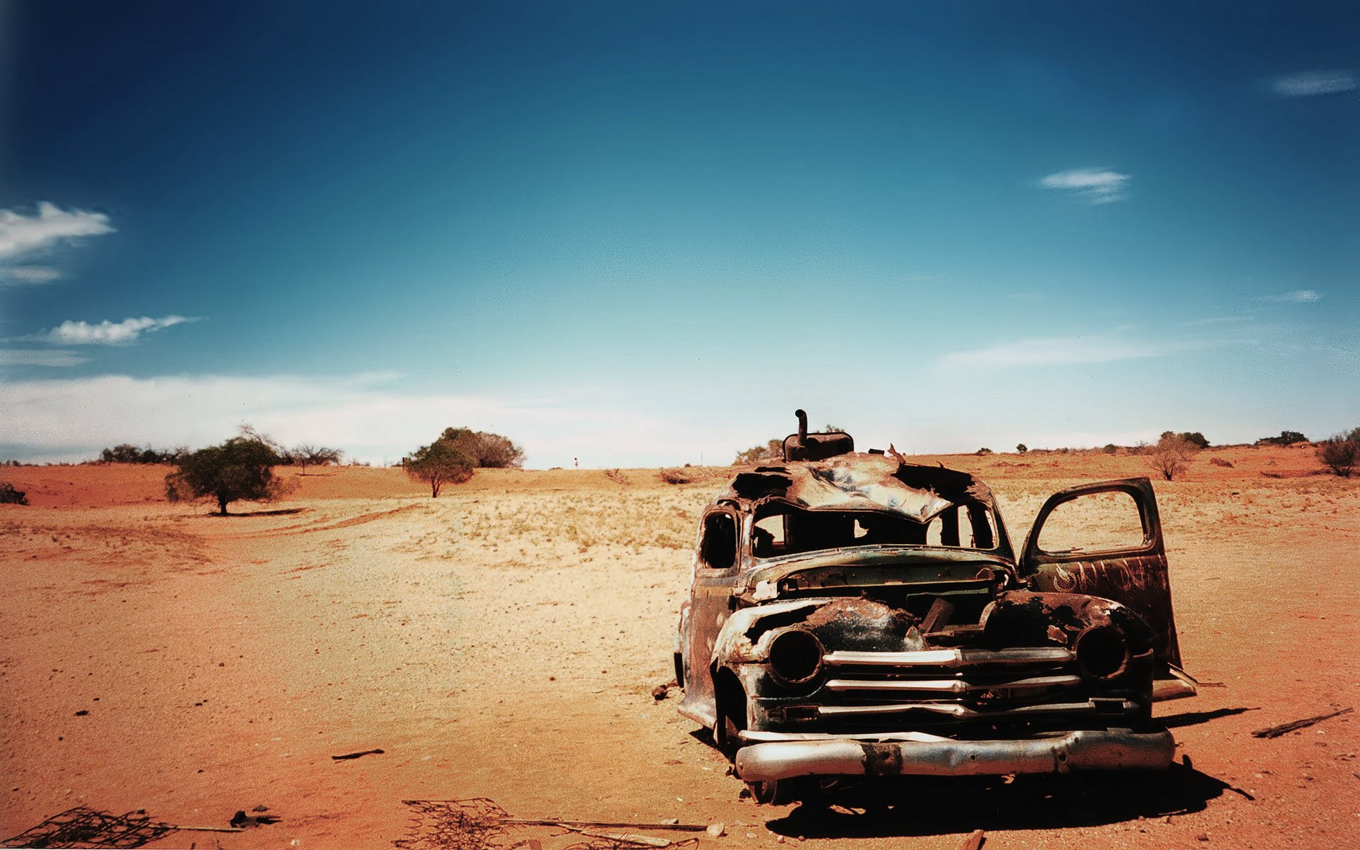 HD desktop wallpaper of a rusted, wrecked vehicle abandoned in a desert landscape under a clear blue sky.