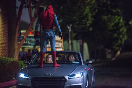 Spider-Man stands on the hood of a silver car at night, confronting two people inside. Scene from the movie Spider-Man: Homecoming. This HD desktop wallpaper and background captures the action and drama.