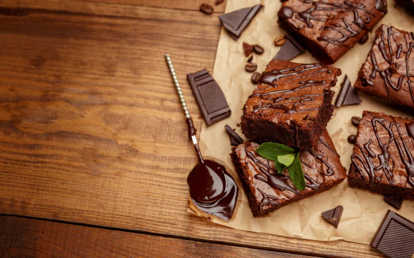 Close-up 4K Ultra HD desktop wallpaper of rich chocolate brownies with chocolate pieces and sauce on parchment paper over a wooden surface.