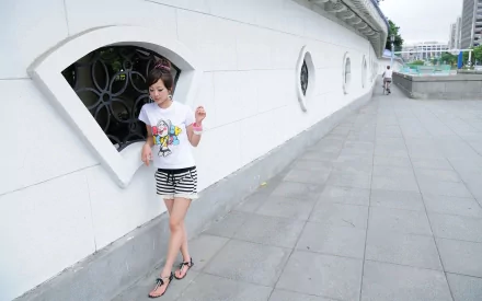 A woman named Mikako Zhang Kaijie leans against a white wall with decorative windows, wearing a colorful t-shirt and striped shorts in this HD desktop wallpaper.