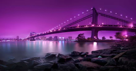 HD desktop wallpaper of the Manhattan Bridge at night in New York City, showcasing a vibrant purple sky reflecting over the river, with city lights in the background and rocks in the foreground.