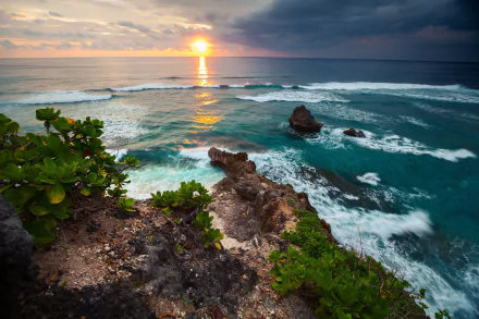 HD desktop wallpaper featuring a breathtaking Indonesian coastline at sunset, with vibrant ocean waves crashing against rugged rocks under a colorful horizon.
