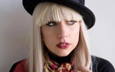 A close-up of Lady Gaga with striking blonde hair and bold makeup, wearing a black hat and colorful scarf, creating a captivating music-themed HD desktop wallpaper.
