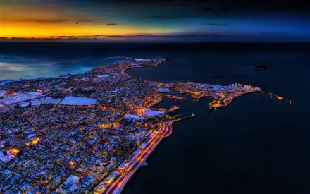 Aerial night view of Reykjavik, Iceland, showcasing city lights along the coastline with a vibrant horizon in this HD desktop wallpaper.