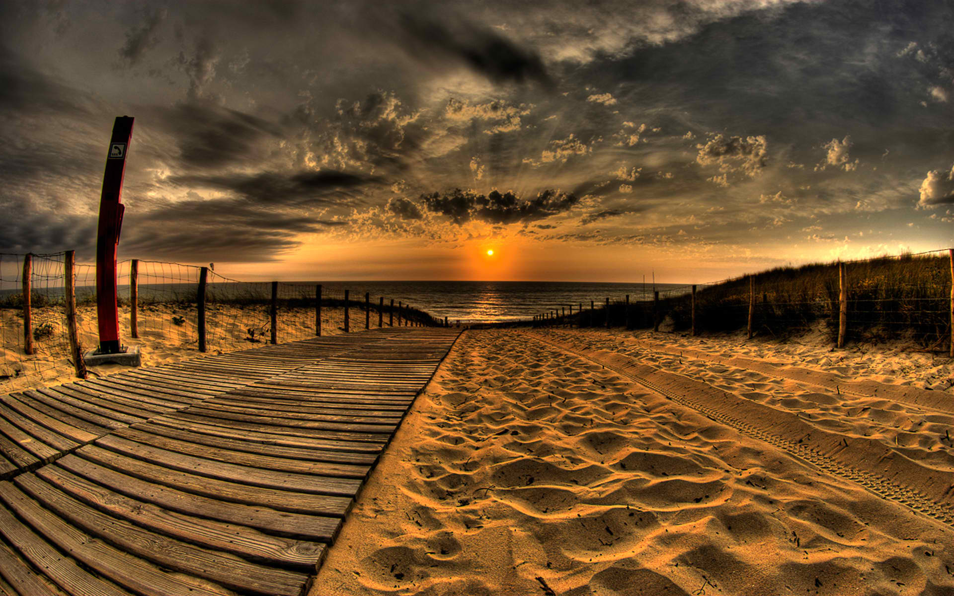 A serene sunset over a sandy beach, with a fence, clouds, water, and the Earth in view.