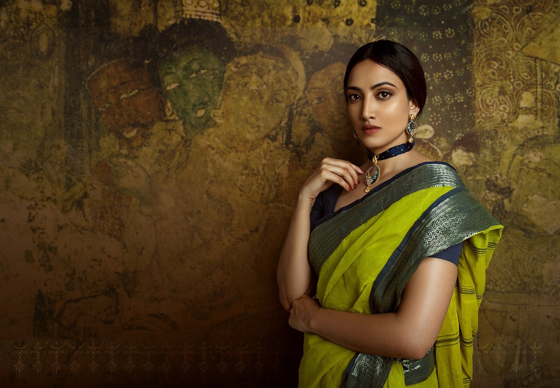 A woman with brown eyes and brunette hair wears earrings, a necklace, and a saree. She stands against a backdrop of Indian oriental art in this HD artistic desktop wallpaper.