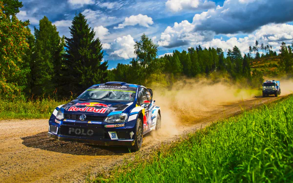 A race car kicks up dust while speeding on a dirt road during a rally race, surrounded by greenery and a vibrant blue sky. This HD desktop wallpaper captures the thrill and excitement of rallying sports.