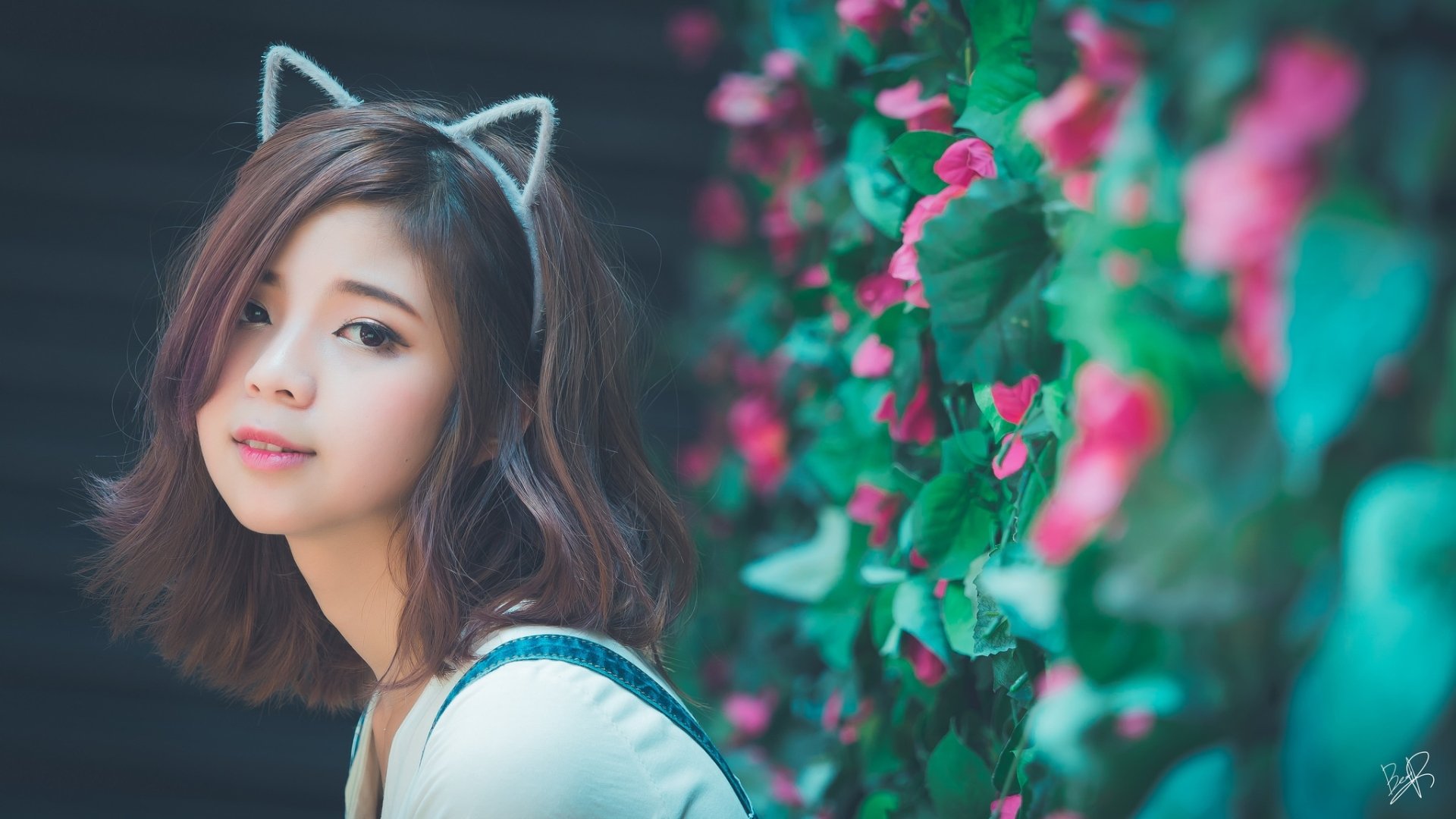 HD wallpaper featuring an Asian brunette model wearing cat ears, standing against a backdrop of vibrant green leaves and pink flowers.