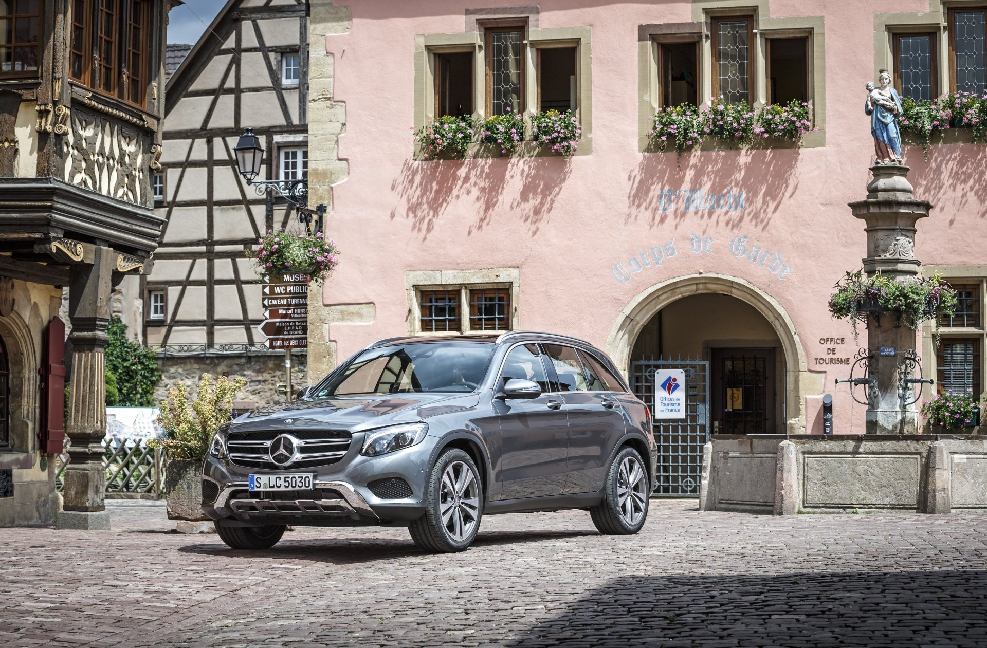 Silver Mercedes-Benz GLC-Class SUV on a cobblestone square before a pink half-timbered building - 4K Ultra HD PC desktop wallpaper of the car.