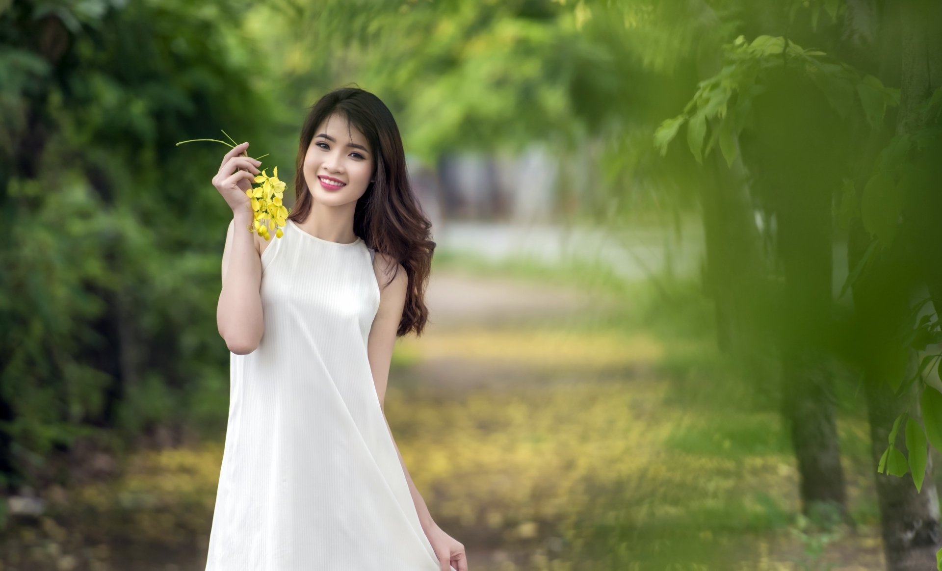 A brunette Asian woman in a white dress smiles while holding yellow flowers against a lush, blurred green background. The image captures her brown eyes and cheerful demeanor, making it a vibrant HD wallpaper.