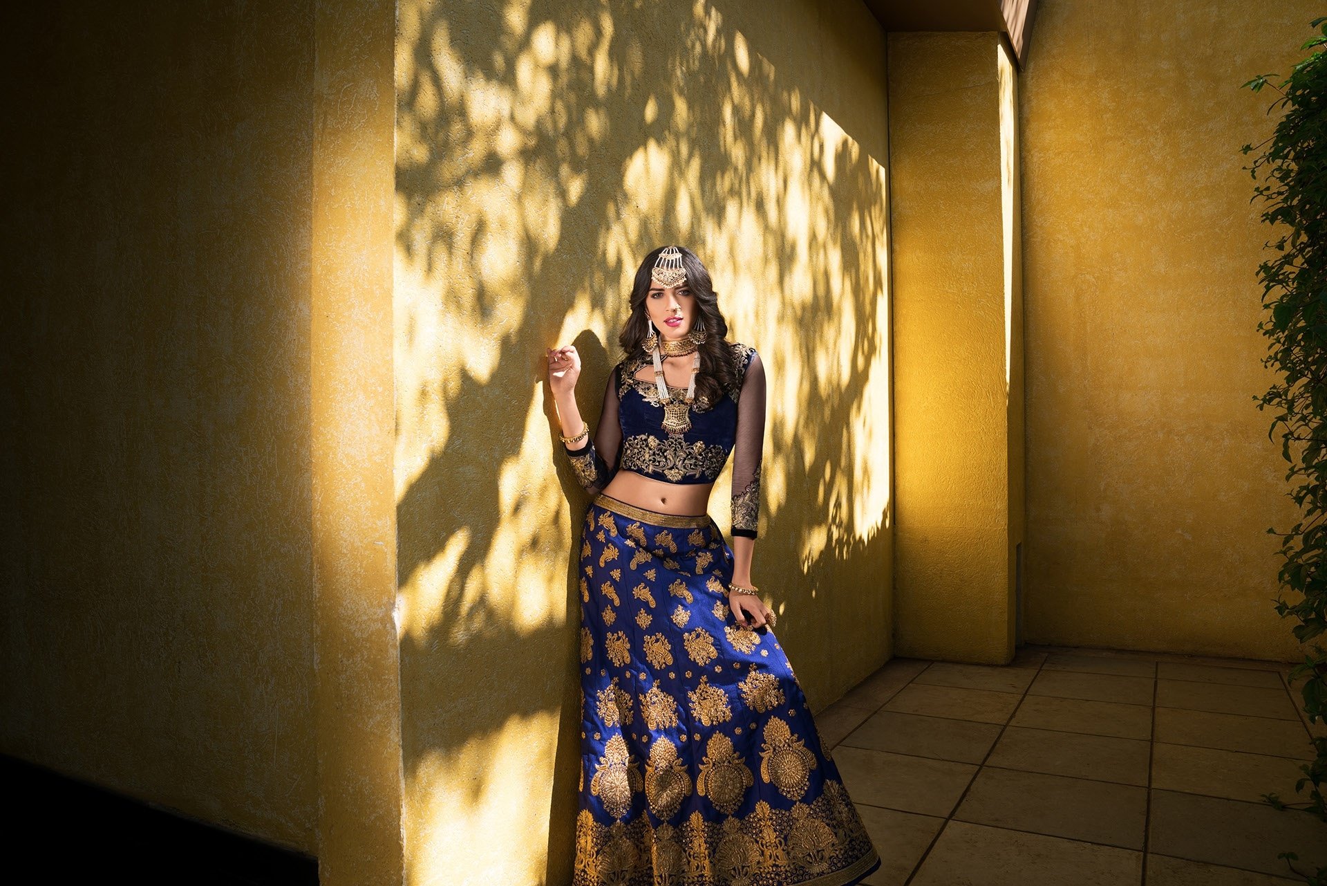 Brunette woman in ornate necklace and jewelry wearing a blue-and-gold saree, posed in a sunlit courtyard — artistic HD PC desktop wallpaper/background.