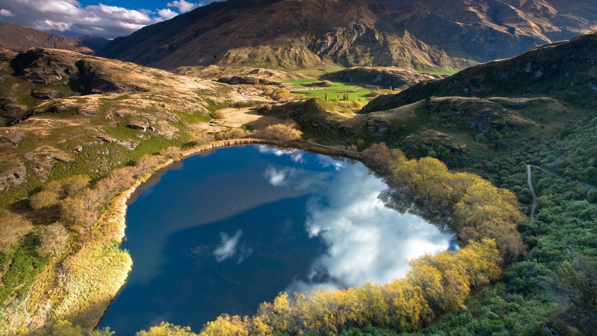A serene lake surrounded by lush trees and rolling hills, reflecting the sky above, captures the beauty of nature in this stunning HD desktop wallpaper.