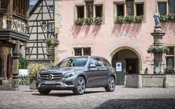 Silver Mercedes-Benz GLC-Class SUV on a cobblestone square before a pink half-timbered building - 4K Ultra HD PC desktop wallpaper of the car.