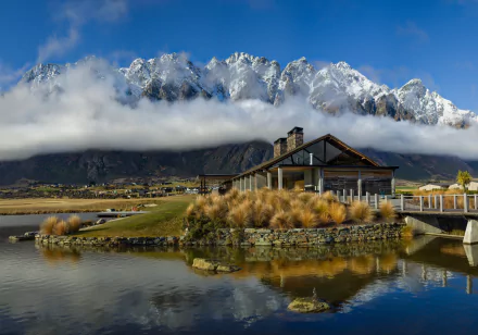 A man-made restaurant by a calm lake with tall grasses, set against New Zealand’s mountain landscape partly covered by clouds, captured in 8K Ultra HD.