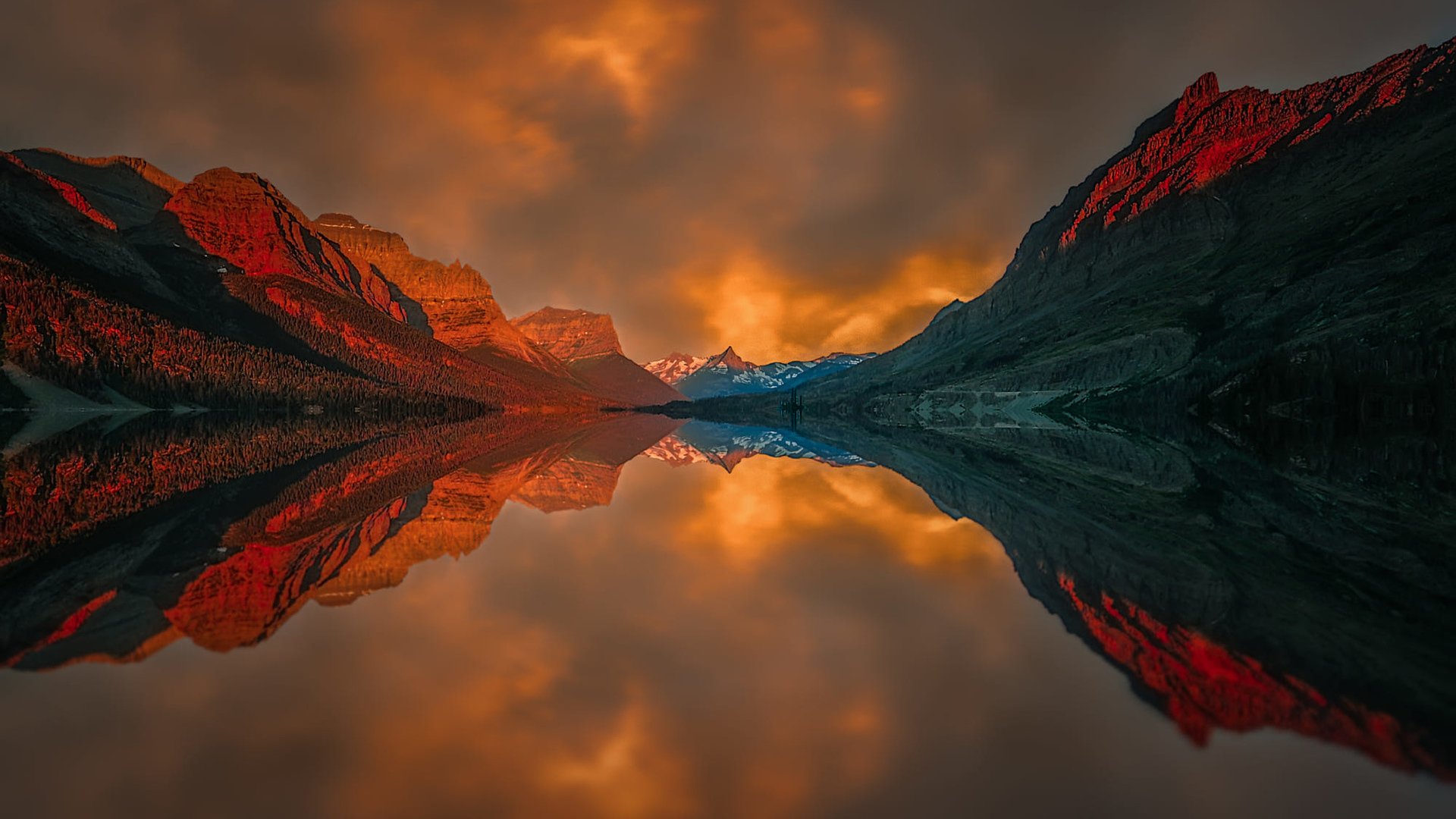 A stunning sunset reflects off a tranquil lake, with vibrant clouds and rugged mountains silhouetted against the dramatic sky. This HD nature wallpaper captures the serene beauty of the outdoors.
