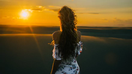 HD PC desktop wallpaper and background showing a woman from the rear with long curly hair standing on sand dunes at sunset, bathed in warm golden light.