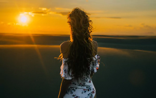 HD PC desktop wallpaper and background showing a woman from the rear with long curly hair standing on sand dunes at sunset, bathed in warm golden light.