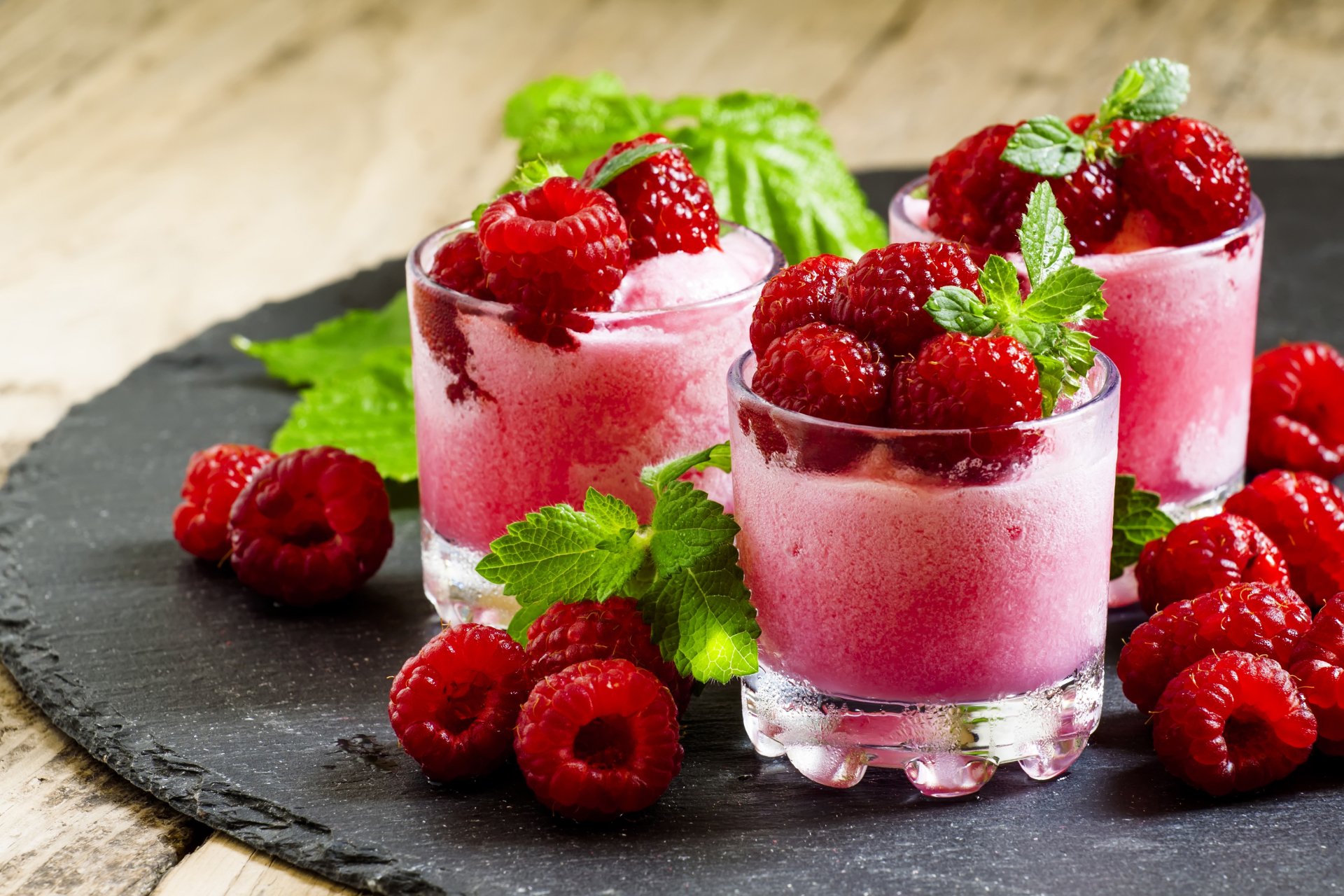 Close-up still life of fresh raspberries and raspberry smoothies in glasses, presented on a dark slate surface, captured in vibrant 4K Ultra HD for a PC desktop wallpaper.