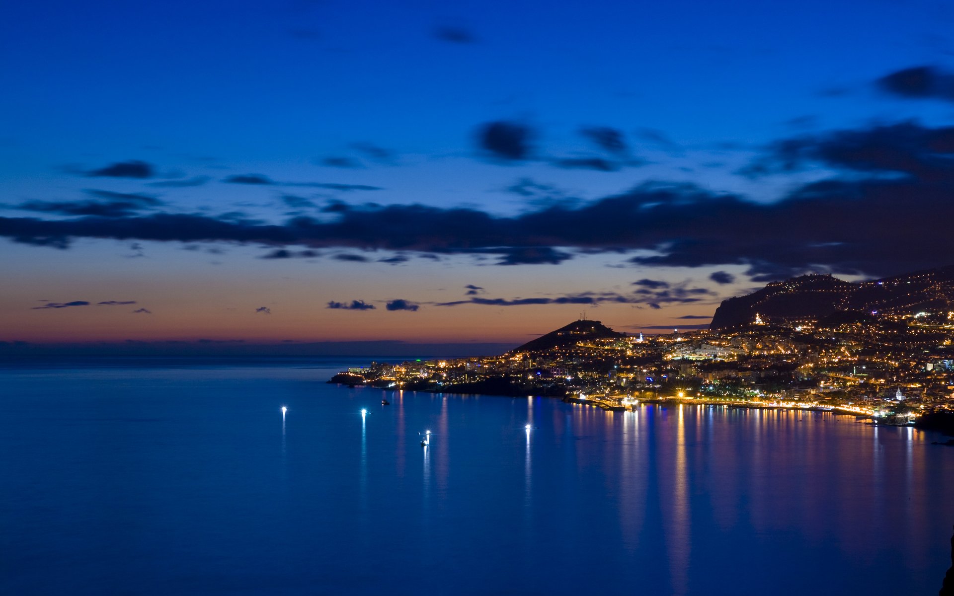 A breathtaking sunset over a city by the ocean, featuring vibrant clouds and shimmering lights reflecting on the water, creating a stunning HD wallpaper for your desktop.
